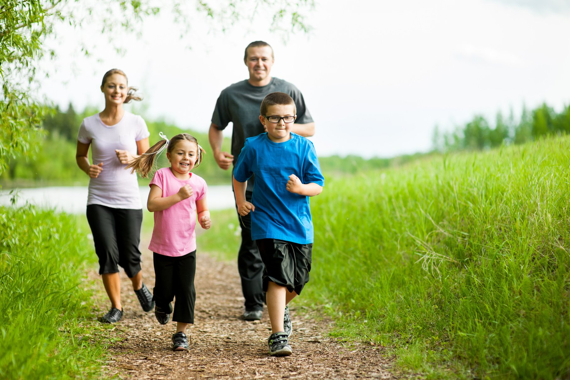 Family enjoying a healthy lifestyle with a jog in a lush green park, featuring two children and parents smiling and exercising together outdoors