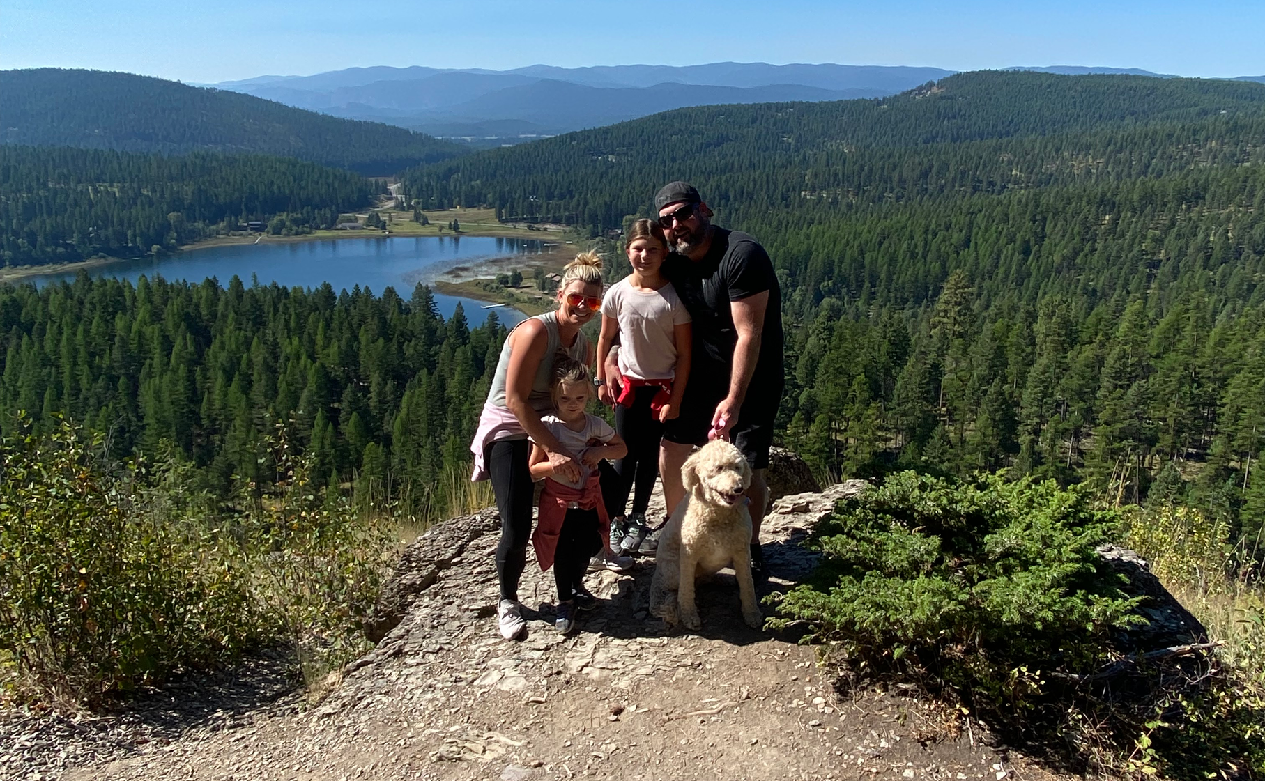 JRP Health coaches Jessica and Randy with their Family and dog enjoying a sunny day hike on a mountain overlook with a scenic lake view in the background, surrounded by dense green forests and distant hills under a clear blue sky